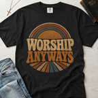 Worship Anyways Christian t-shirt in black, garment-dyed retro graphic tee with vintage 70s sunburst typography in orange and brown tones, styled flat lay with denim.