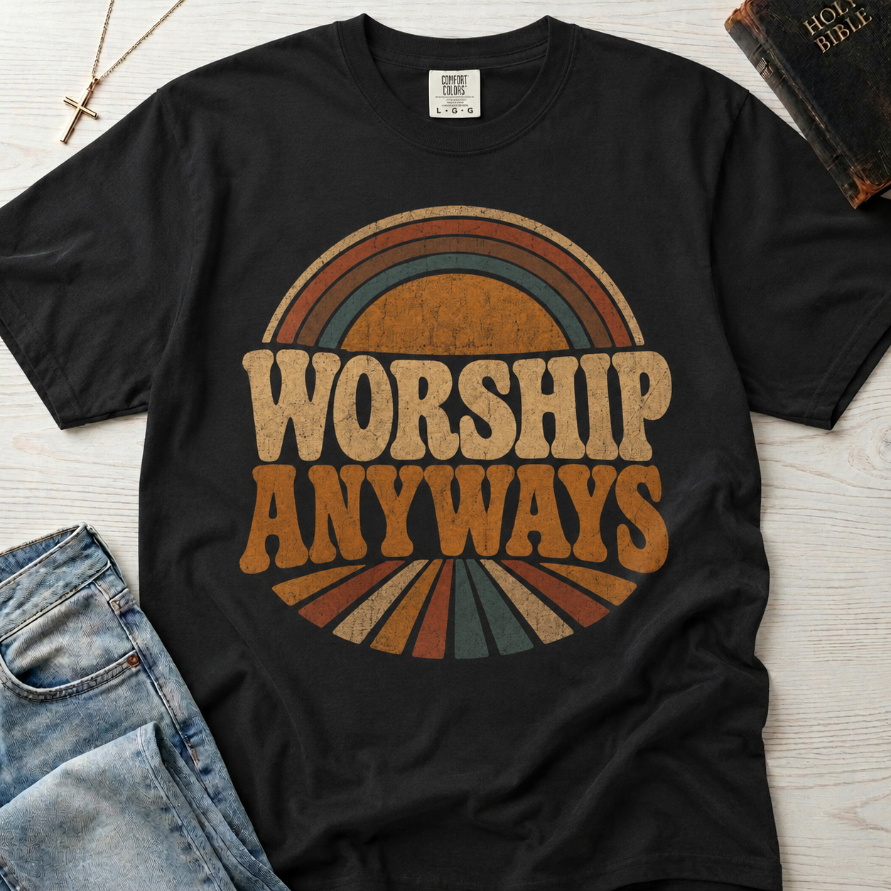 Worship Anyways Christian t-shirt in black, garment-dyed retro graphic tee with vintage 70s sunburst typography in orange and brown tones, styled flat lay with denim.