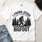 I Found Jesus Still Looking for Bigfoot Christian humor t-shirt in white, outdoor graphic with pine tree silhouettes and Bigfoot illustration.