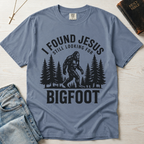 I Found Jesus Still Looking for Bigfoot Christian humor t-shirt in dusty Blue Jean, outdoor sasquatch graphic with pine tree silhouettes.