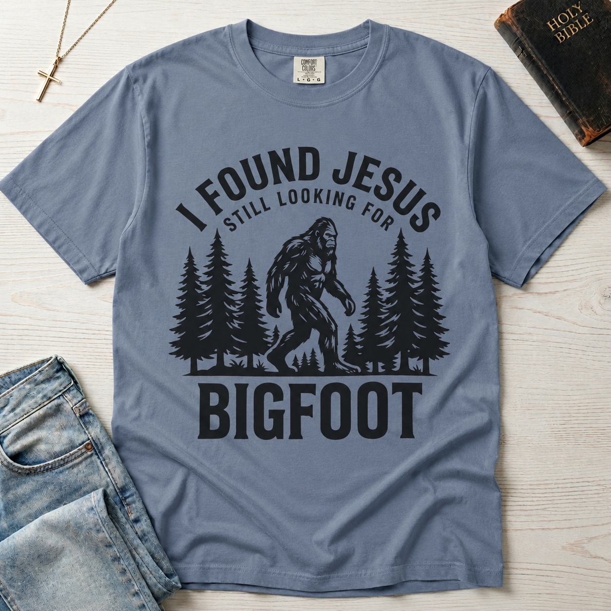 I Found Jesus Still Looking for Bigfoot Christian humor t-shirt in dusty Blue Jean, outdoor sasquatch graphic with pine tree silhouettes.
