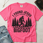 I Found Jesus Still Looking for Bigfoot Christian humor t-shirt in bright Crunchberry pink, outdoor Bigfoot graphic with pine tree silhouettes.