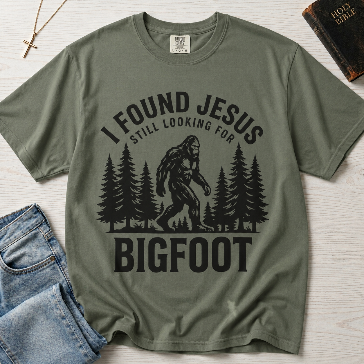 I Found Jesus Still Looking for Bigfoot Christian humor t-shirt in Moss sage green, outdoor design with Bigfoot and pine trees.