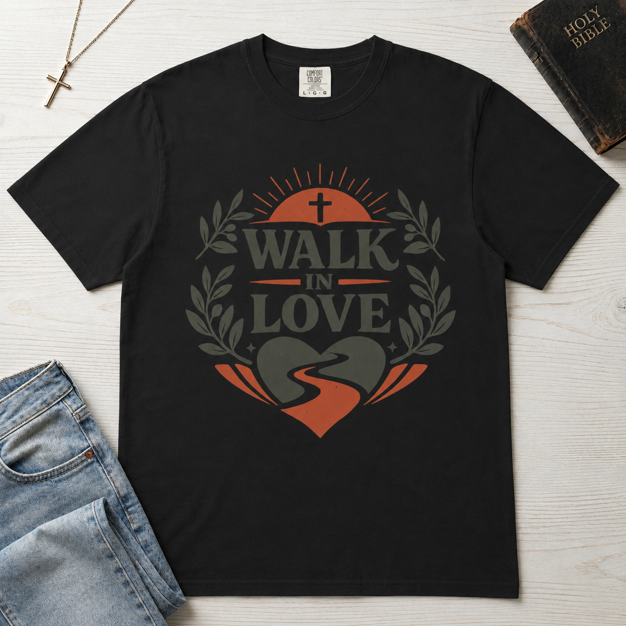 Walk in Love Christian t-shirt in deep graphite black, garment-dyed faith tee with cross and laurel wreath, styled flat lay with Holy Bible and cross necklace.