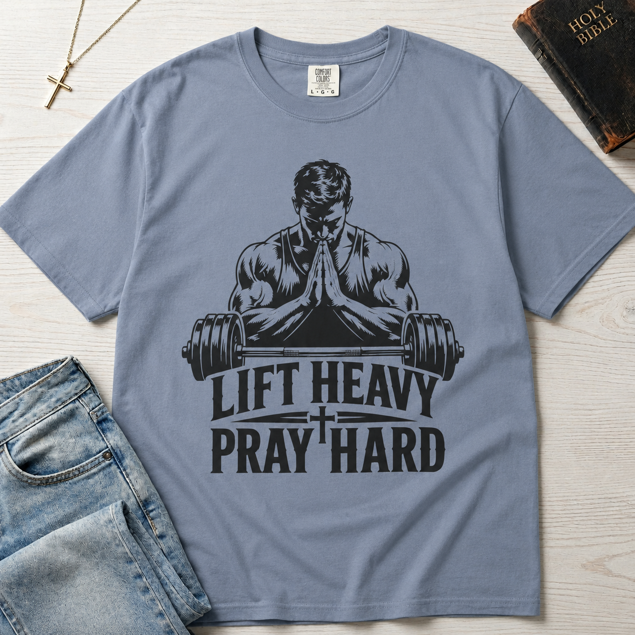 Lift Heavy Pray Hard Christian gym t-shirt in dusty Blue Jean, faith and fitness design with praying weightlifter over barbell, styled flat lay with denim.