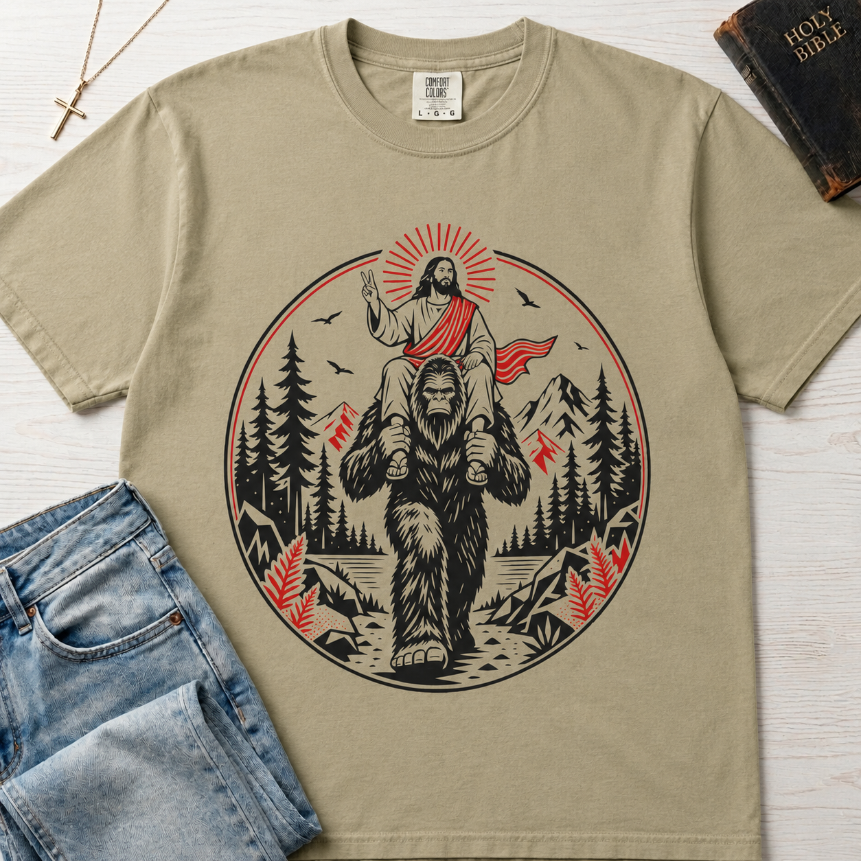 Bigfoot & Jesus Christian t-shirt in tan sand; circular forest emblem of Jesus riding on Bigfoot's shoulders among pines with a sunburst halo.
