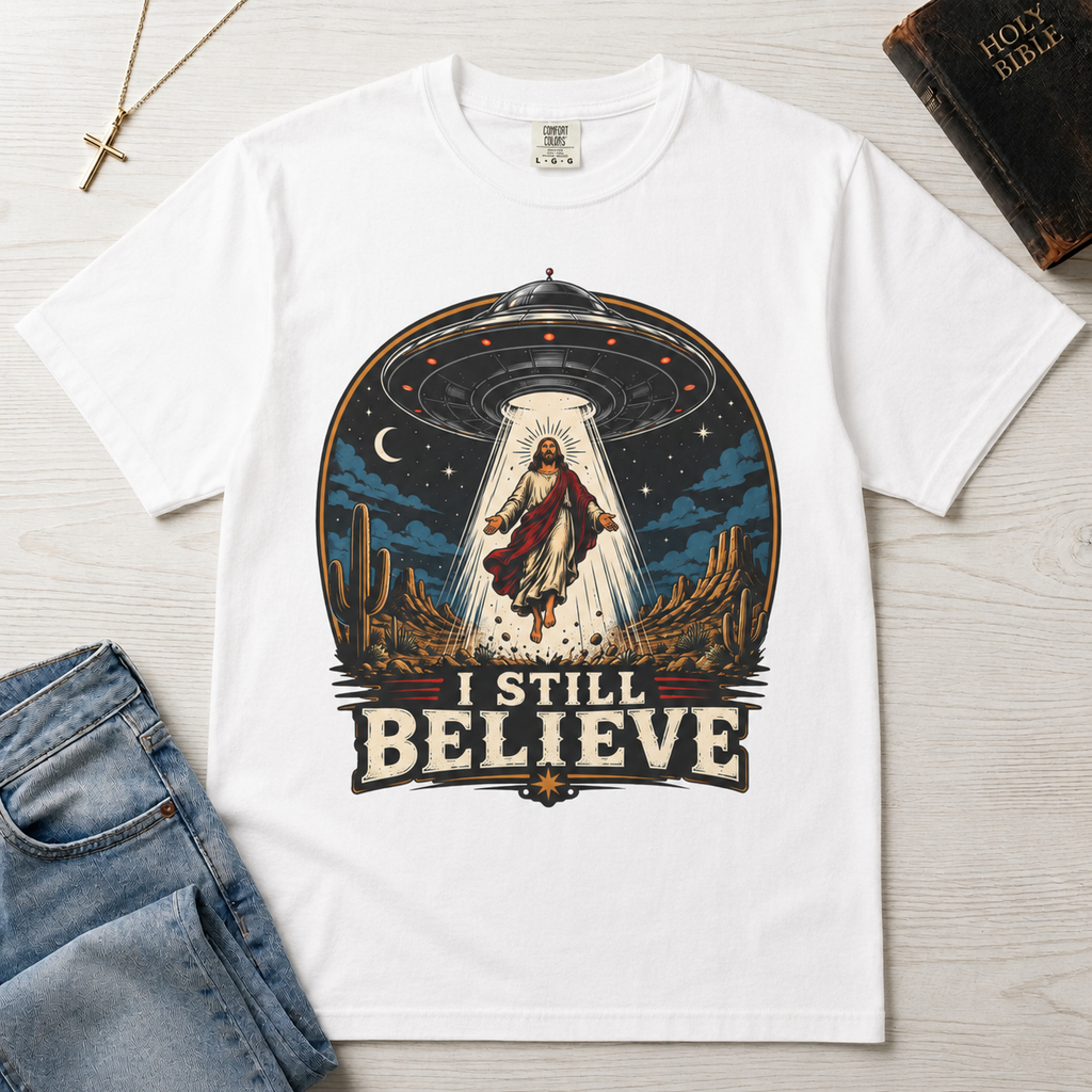 I Still Believe Christian t-shirt in white; retro graphic of a UFO beaming up a figure over desert cactus at night with 'I Still Believe' banner.