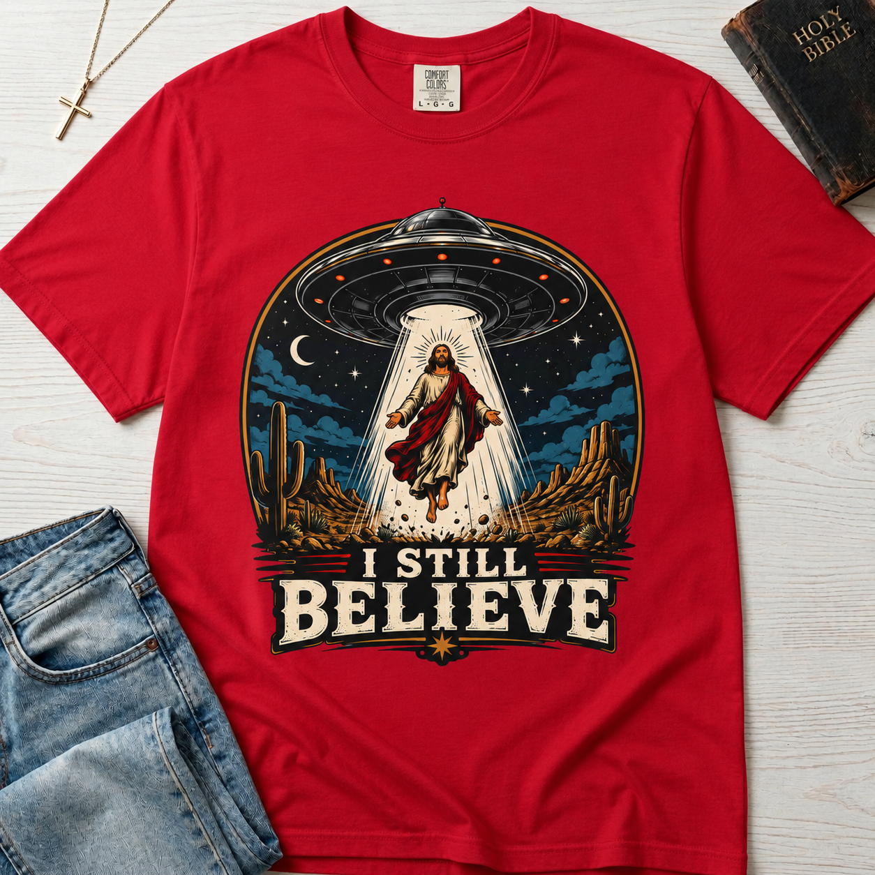 I Still Believe Christian t-shirt in coral red; retro graphic of a UFO beaming up a figure over desert cactus at night with 'I Still Believe' banner.