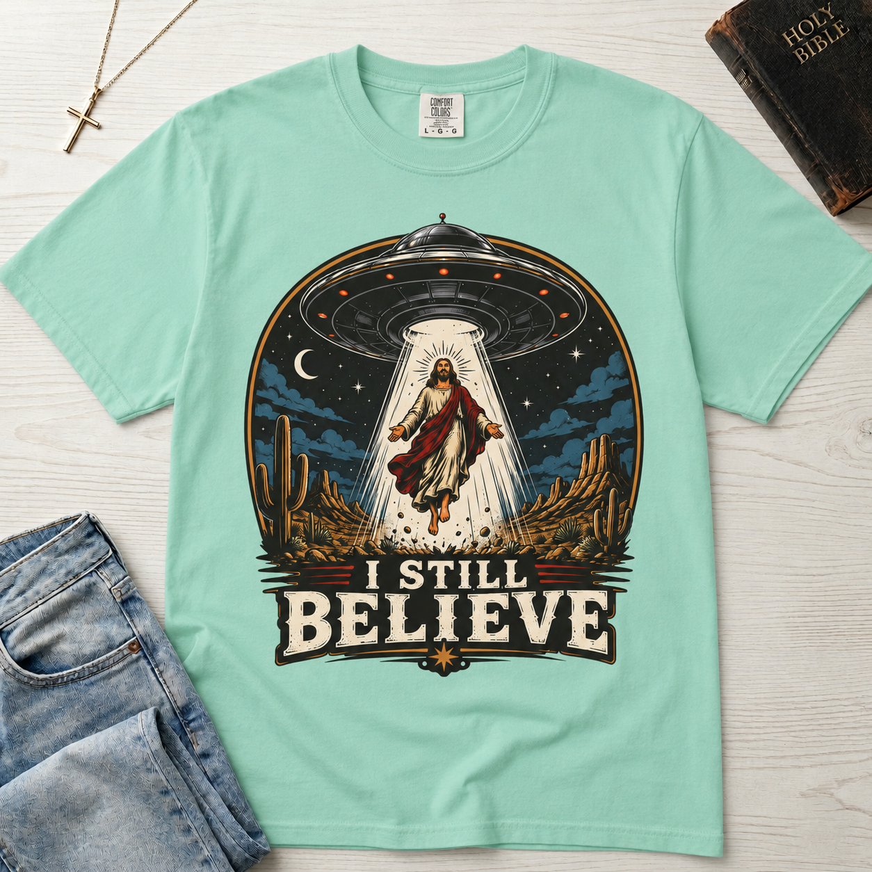 I Still Believe Christian t-shirt in mint green; retro graphic of a UFO beaming up a figure over desert cactus at night with 'I Still Believe' banner.