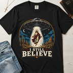 I Still Believe Christian t-shirt in graphite gray; retro graphic of a UFO beaming up a figure over desert cactus at night with 'I Still Believe' banner.