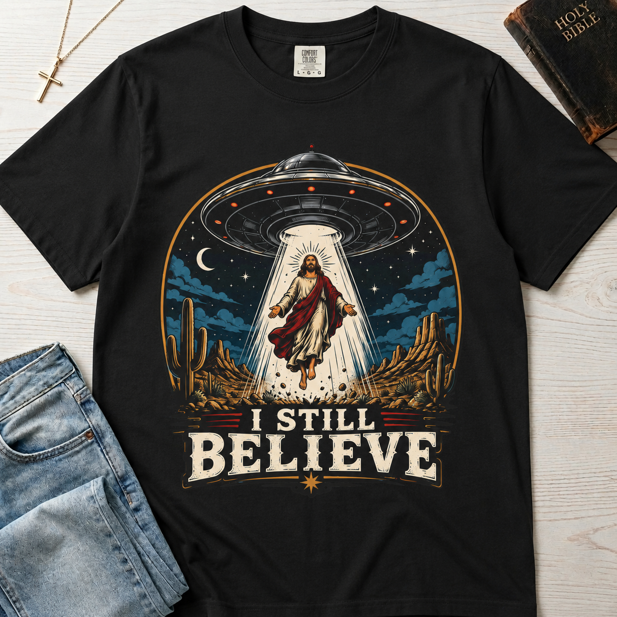 I Still Believe Christian t-shirt in graphite gray; retro graphic of a UFO beaming up a figure over desert cactus at night with 'I Still Believe' banner.