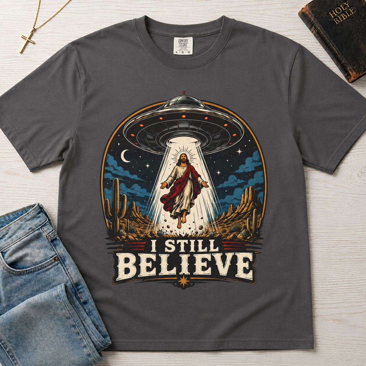 I Still Believe Christian t-shirt in gray; retro graphic of a UFO beaming up a figure over desert cactus at night with 'I Still Believe' banner.