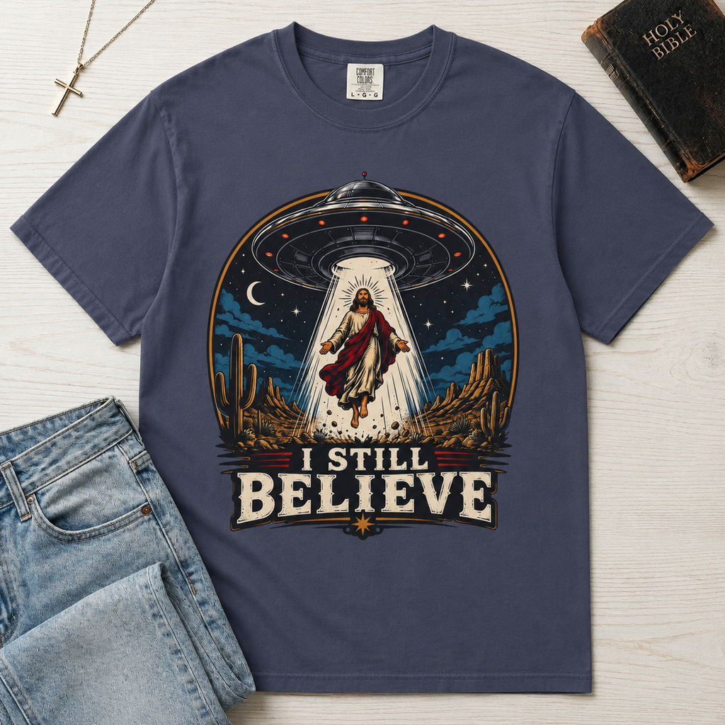 I Still Believe Christian t-shirt in slate blue; retro graphic of a UFO beaming up a figure over desert cactus at night with 'I Still Believe' banner.