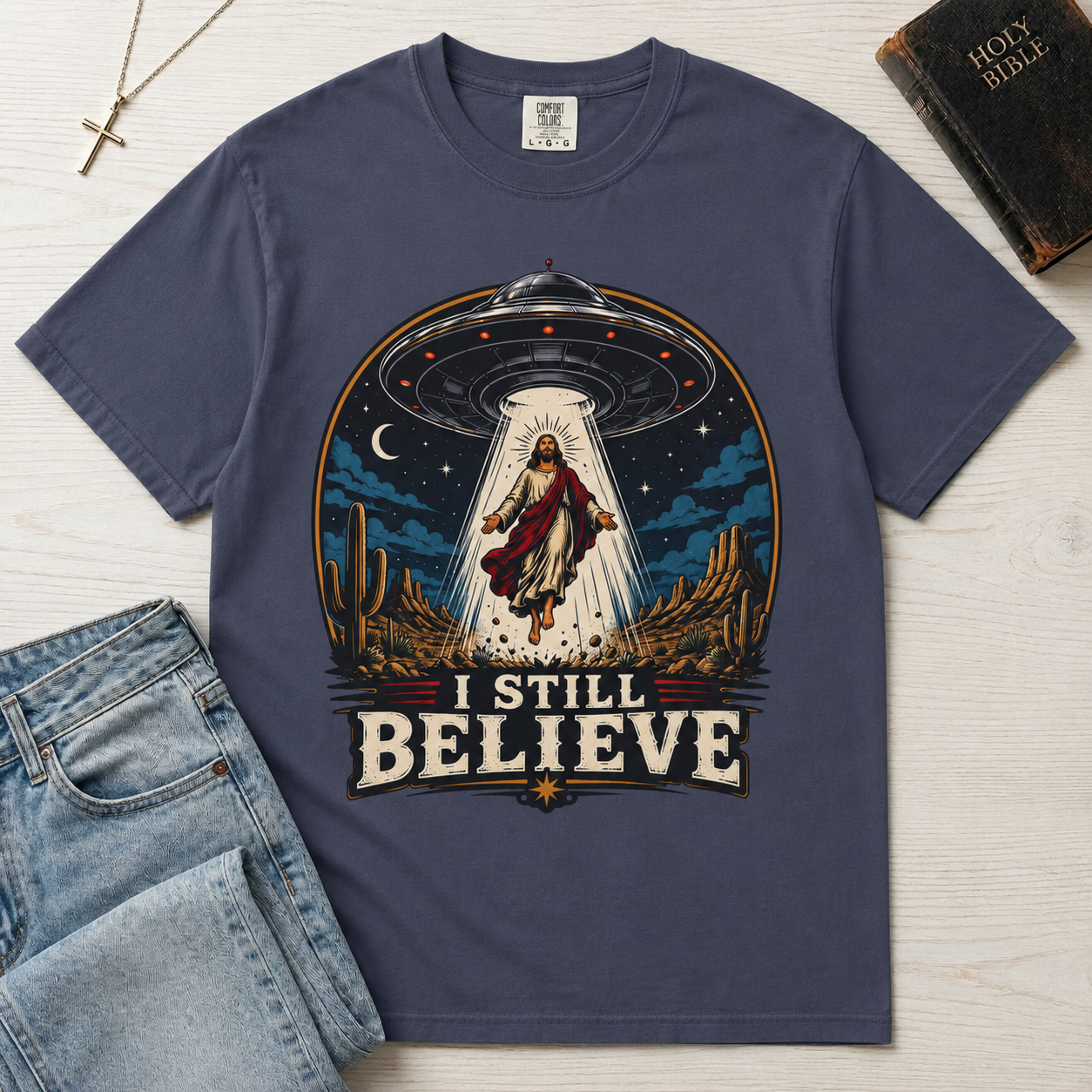 I Still Believe Christian t-shirt in slate blue; retro graphic of a UFO beaming up a figure over desert cactus at night with 'I Still Believe' banner.