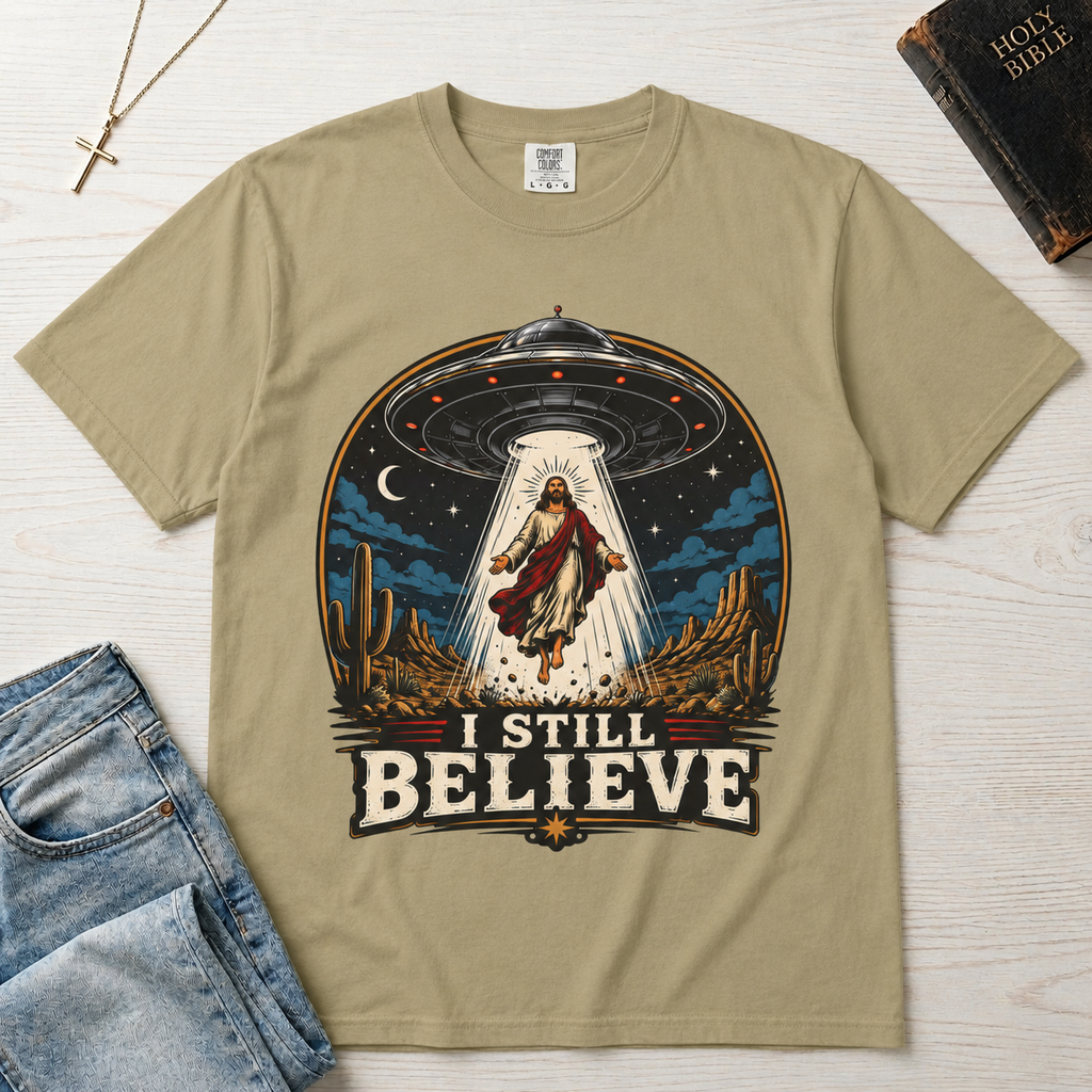 I Still Believe Christian t-shirt in tan sand; retro graphic of a UFO beaming up a figure over desert cactus at night with 'I Still Believe' banner.