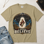 I Still Believe Christian t-shirt in tan sand; retro graphic of a UFO beaming up a figure over desert cactus at night with 'I Still Believe' banner.