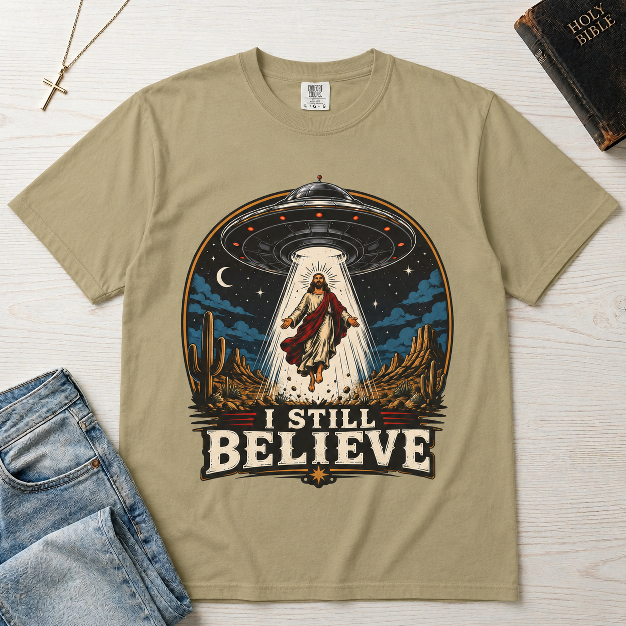 I Still Believe Christian t-shirt in tan sand; retro graphic of a UFO beaming up a figure over desert cactus at night with 'I Still Believe' banner.